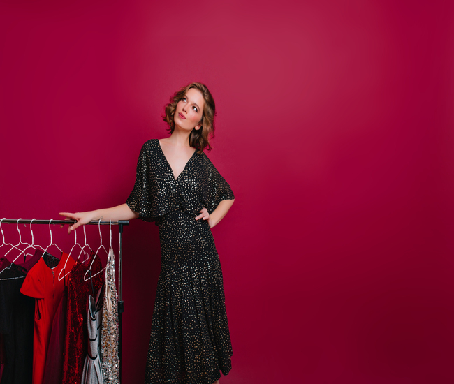 Dreamy woman in retro black dress looking up while posing beside hangers with clothes. Romantic lady with short hairstyle standing in wardrobe and thinking what to wear..